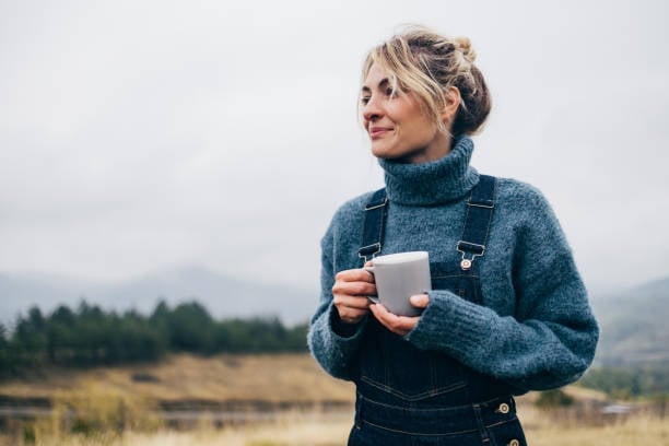 Smiling woman in a teal sweater holding a white mug outdoors, with mountains and forest in the background, in a quiet moment of understanding her own body and cycle