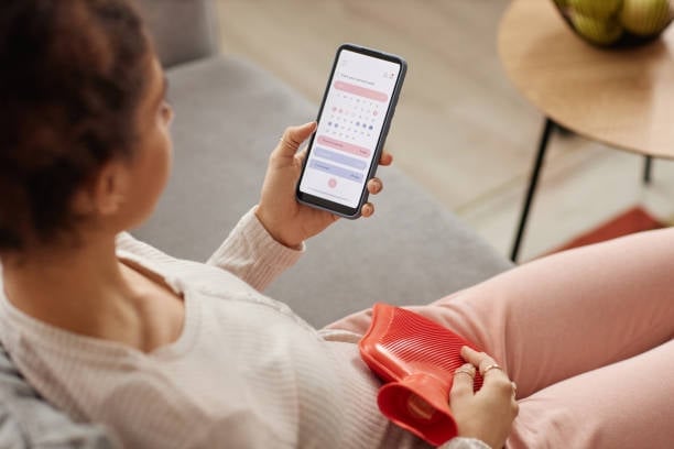 Woman holding a smartphone showing a calendar app interface, with a hot water bottle nearby, seated indoors in casual cream clothing