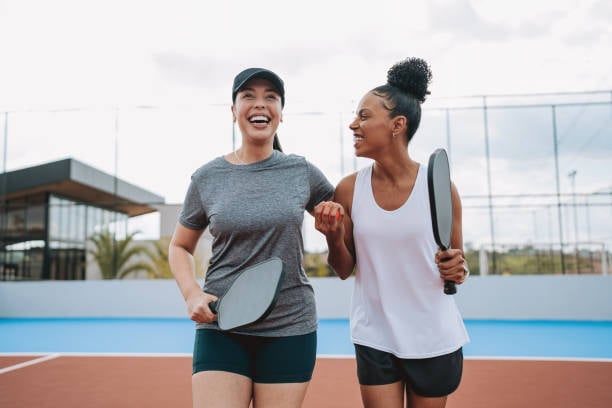 Two female athletes laughing together on an outdoor tennis court on a sunny day, holding pickleball paddles