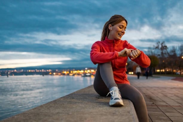 Smiling woman in a red jacket sitting by a waterfront at dusk checking her smartwatch, the way cycle tracking often starts