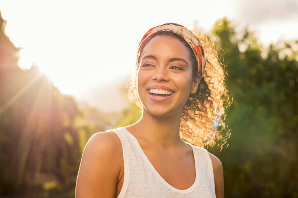 Smiling woman wearing a white tank top and a headband in a sunlit outdoor setting with green trees in the background